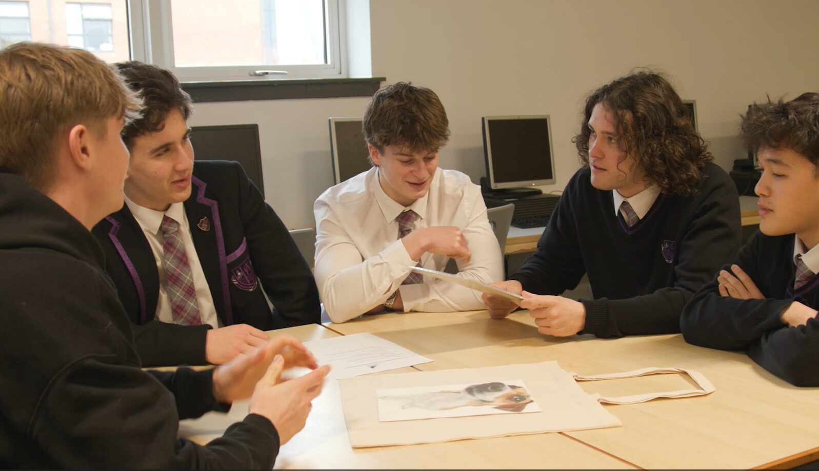 A group of Clydeview Academy pupils in uniform sit around a table in a classroom, discussing and reviewing printed materials together, with one student holding up a sheet while others listen and contribute.