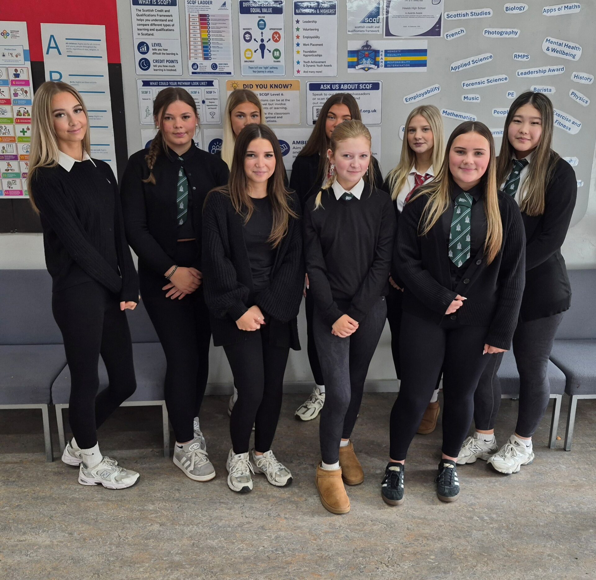 Group photo of nine Hawick High School students in school uniform standing in front of an information display board covered with posters about the Scottish Credit and Qualifications Framework (SCQF), employability, and subject choices. Several students wear green school ties, and the group is arranged in two rows in a school common area with seating visible along the wall behind them.