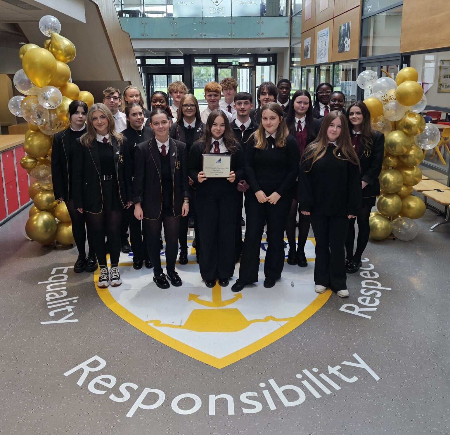 Group of approximately 20 secondary school pupils in school uniform standing in the atrium of Inverclyde Academy, framed by gold and white balloon displays. A pupil at the front holds a plaque, while the group stands on the school crest surrounded by the words “Equality,” “Respect,” and “Responsibility.” The bright, modern school interior is visible in the background.
