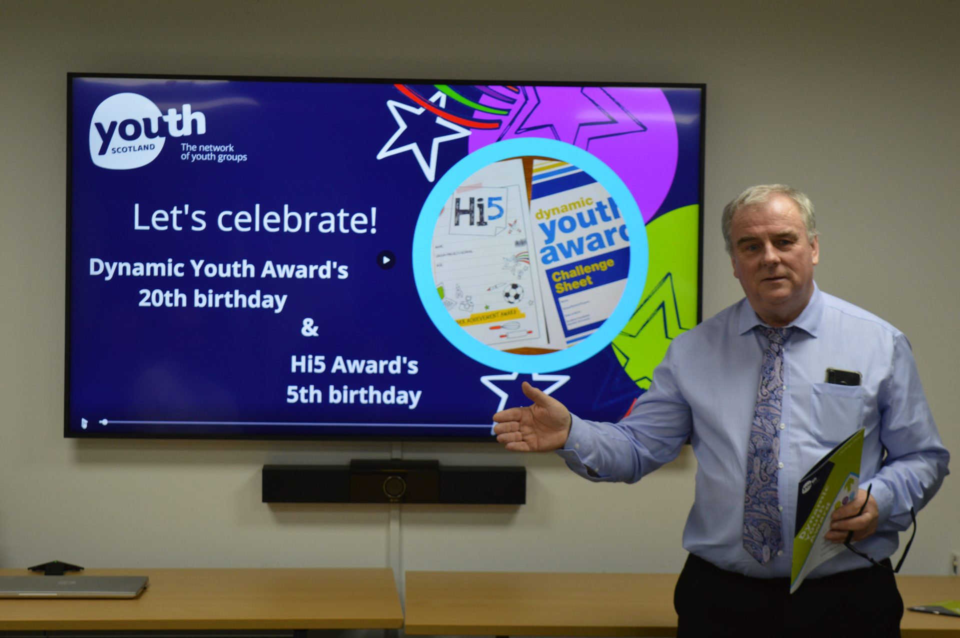 George Cherrie standing beside a large presentation screen displaying a Youth Scotland slide celebrating the Dynamic Youth Award’s 20th birthday and the Hi5 Award’s 5th birthday. The screen features colourful graphics, award materials and Youth Scotland branding. The person gestures towards the display while holding a folder in a meeting room.