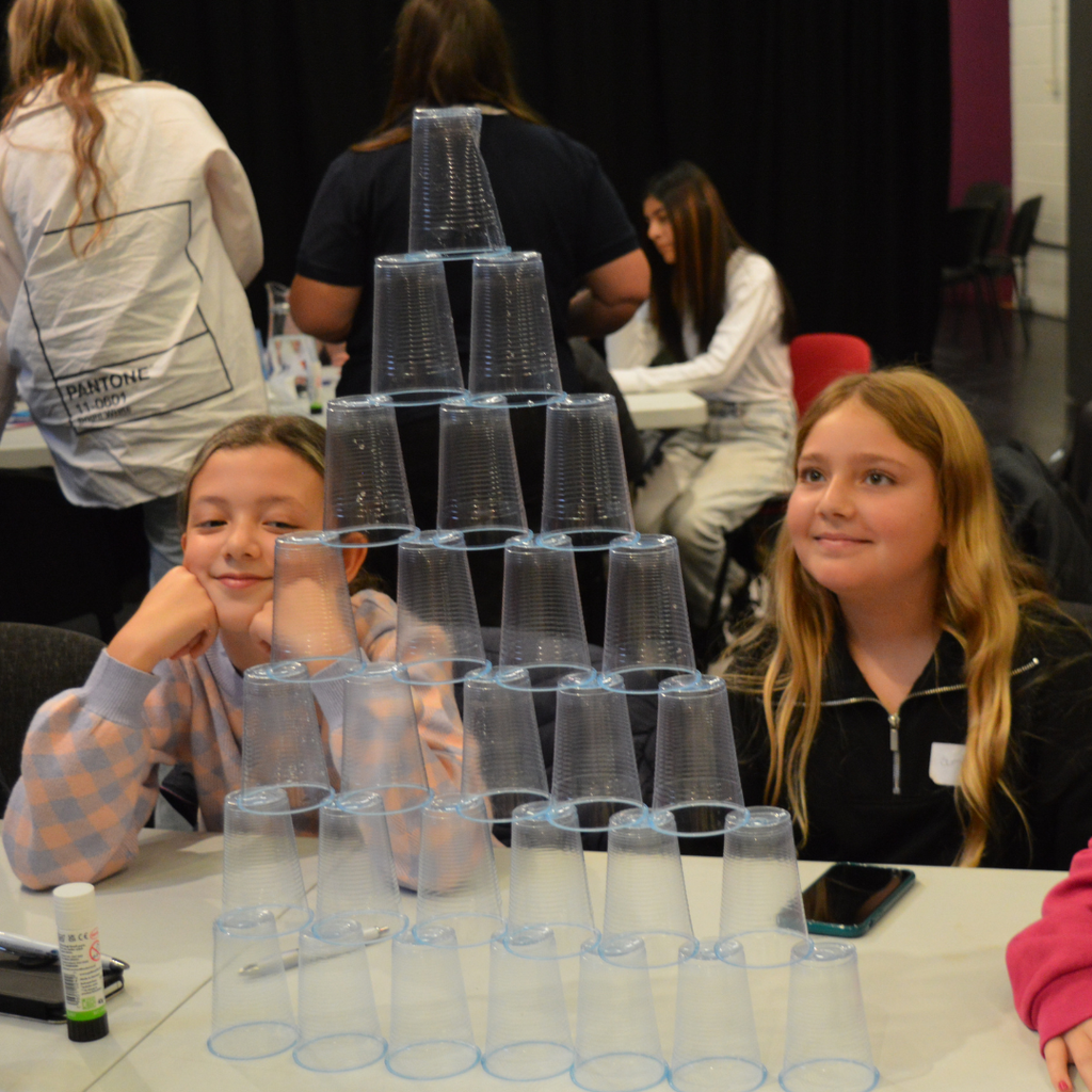 Two young participants sit at a table indoors beside a tall pyramid structure built from clear plastic cups during a group activity. Other participants can be seen working at tables in the background. The cup tower, arranged in a symmetrical triangular shape, is the focal point of the image, highlighting teamwork, problem-solving and creative challenge activities.
