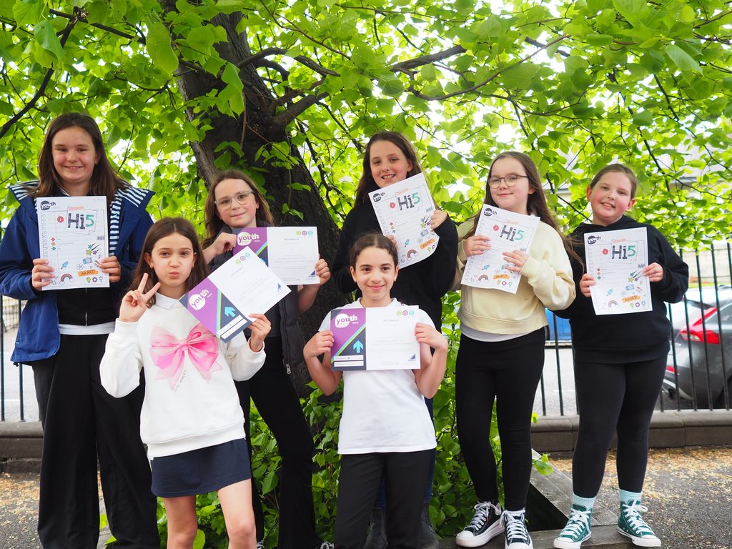 Six young people standing outdoors beneath a large leafy tree, proudly holding Hi5 Award booklets and certificates from Youth Scotland. The group poses together in front of the tree, with several participants displaying completed award materials. The bright green foliage creates a vibrant backdrop, highlighting the celebration of their achievements.